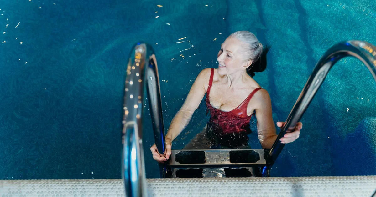 Smiling older woman climbing out of a swimming pool, holding the rails with both arms